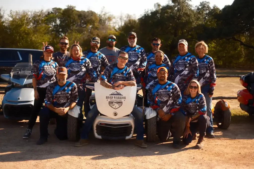 A group of people wearing Road Warrior Foundation long-sleeved shirts, supported by Kraken Bookkeeping, pose with three-wheeled motorcycles outdoors.