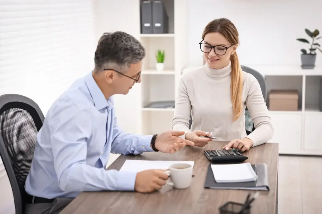 A man and a woman in an office sit across from each other at a desk discussing paperwork and using a calculator for bookkeeping cleanup services.