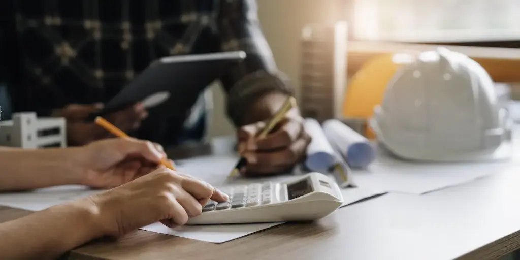 Two people performing construction bookkeeping at a table with blueprints, a calculator, and a hard hat.