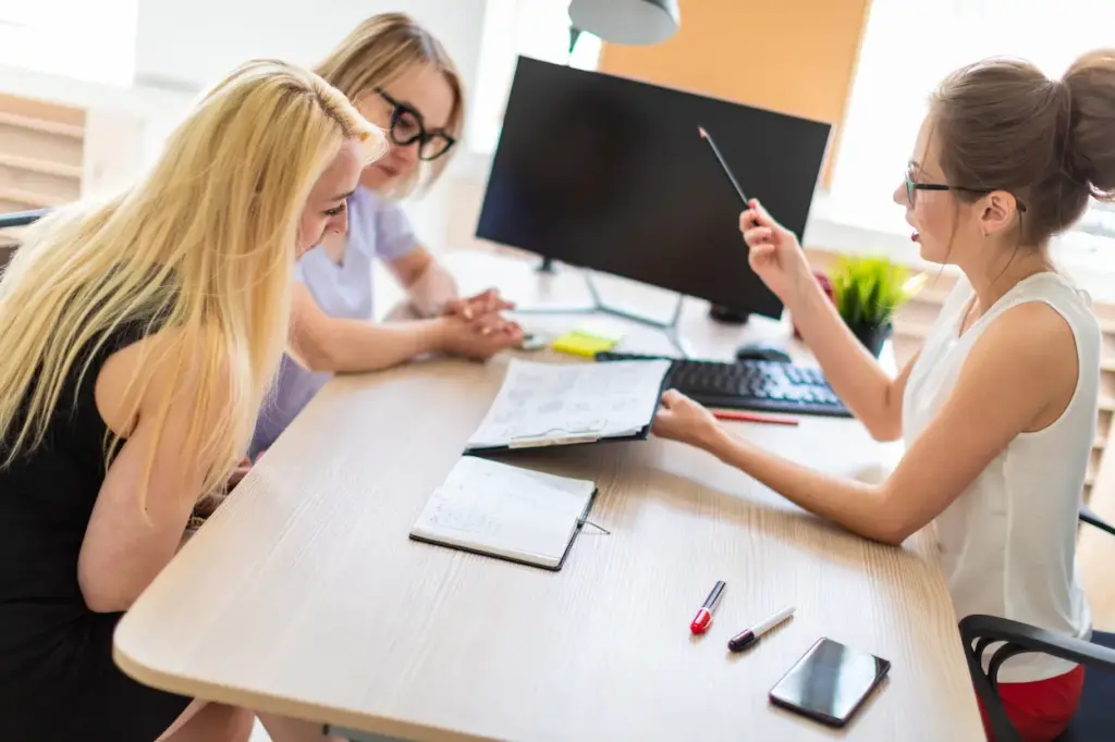 Three women sit at a desk during a monthly bookkeeping meeting, with one woman gesturing toward a document while the others look on.