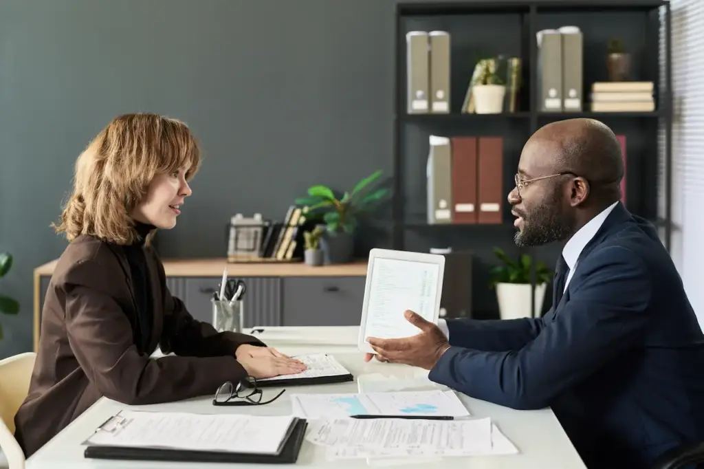 A man in a suit shows a digital tablet to a woman across a desk while discussing bookkeeping cleanup services in an office.