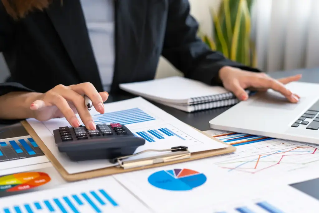 A person in a blazer uses a calculator and a laptop while managing construction bookkeeping with various financial charts and reports on a desk.