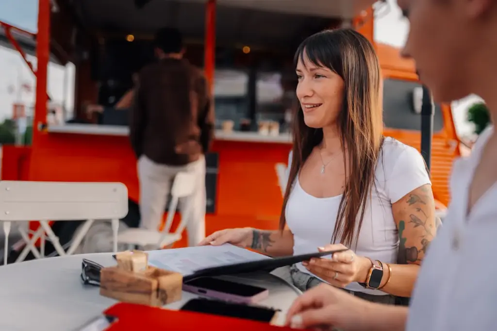 A woman with long dark hair and tattooed arms sits at an outdoor table with a companion, reviewing food truck bookkeeping documents while facing away from a bright orange food truck.