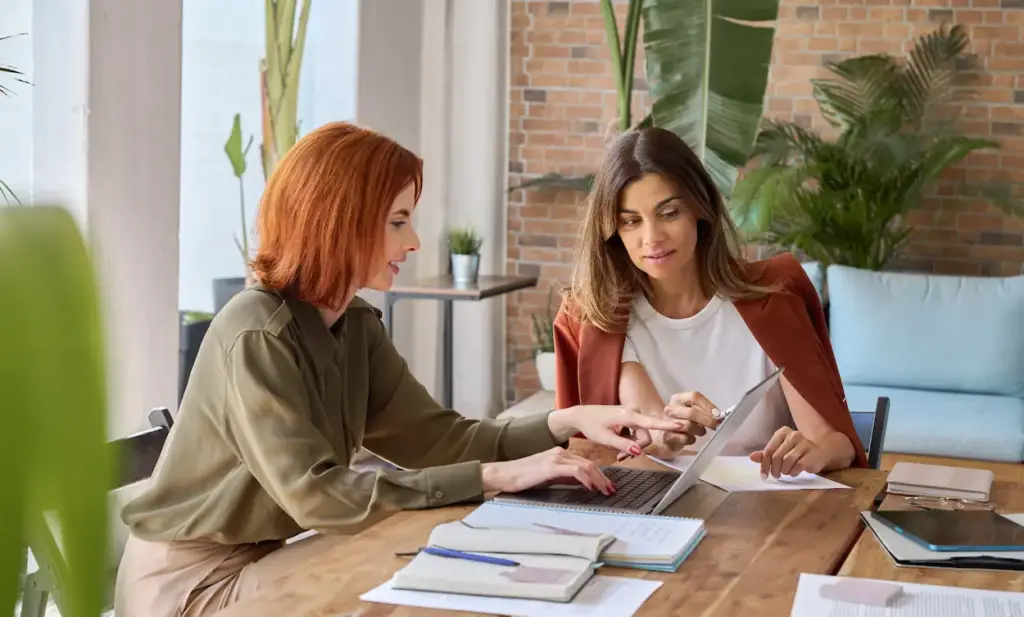 Two women sit at a wooden table reviewing bookkeeping services in Kissimmee on a laptop.