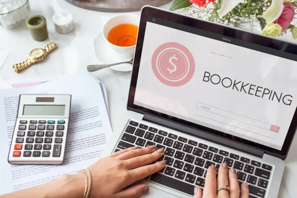 A person typing on a laptop that displays a website titled “BOOKKEEPING” for monthly bookkeeping, next to a calculator and paperwork.