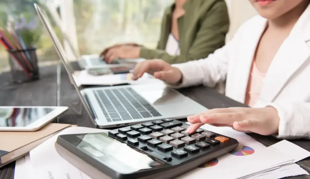 A person performing monthly bookkeeping with a calculator at a desk with a laptop and documents, while another person works on a laptop in the background.