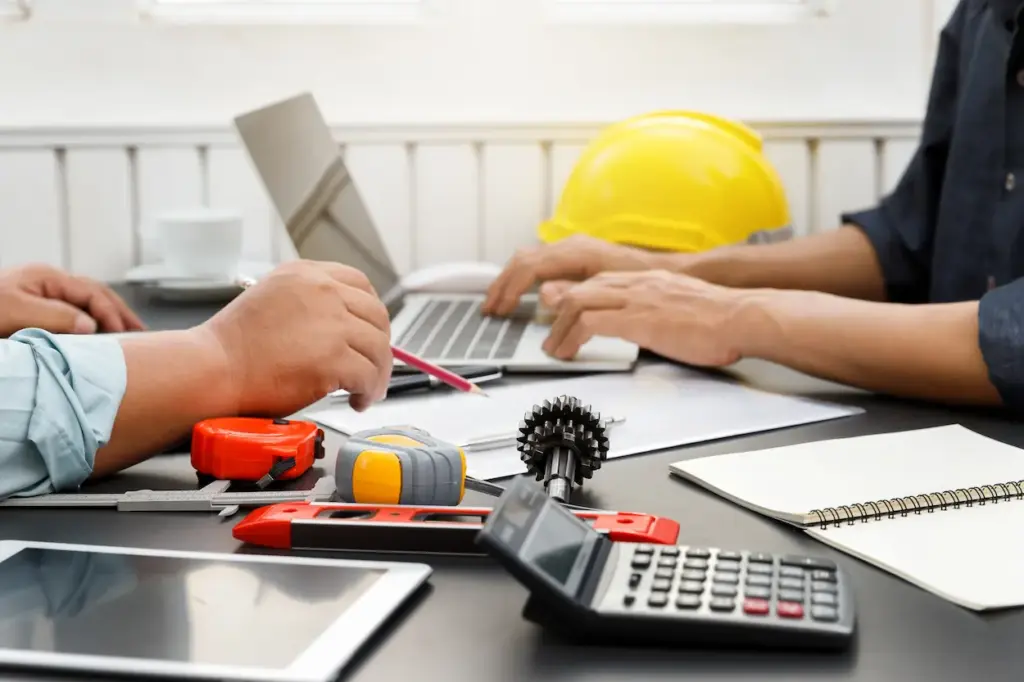 Two people collaborating at a desk with a laptop, a yellow hard hat, measuring tools, a gear, and a calculator to manage construction bookkeeping.