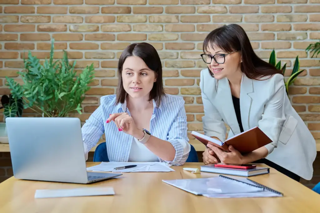 Two women collaborate on QuickBooks training at a desk with a laptop and documents in front of a brick wall.