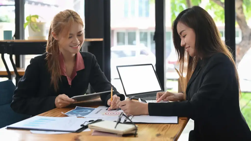 Two businesswomen sitting at a wooden desk reviewing financial documents for a professional bookkeeping service while using a tablet and calculator.