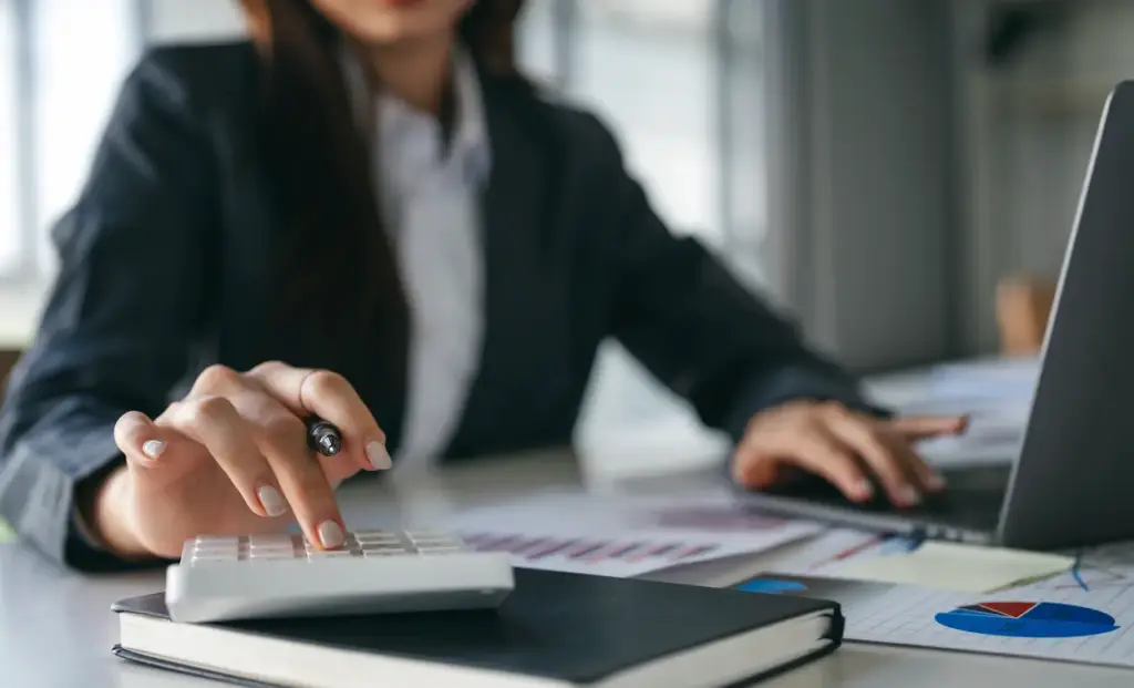 A person in a business suit uses a calculator at a desk with financial charts and a laptop while providing a bookkeeping service in Tampa.