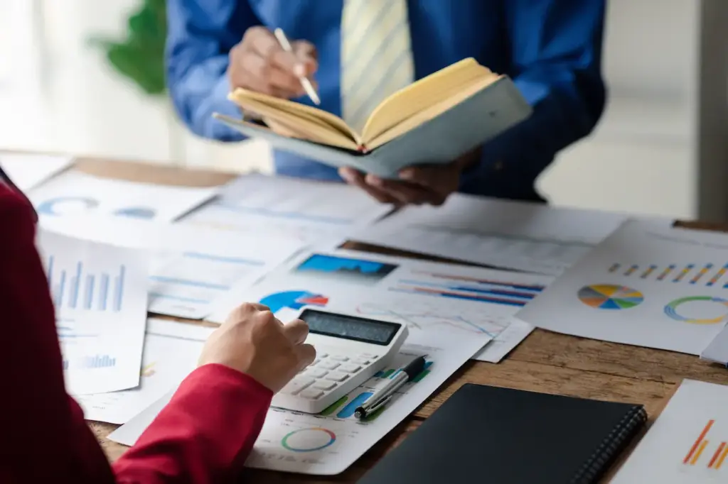 Two professionals reviewing financial documents and charts on a wooden desk, one using a calculator and the other taking notes in a planner for a bookkeeping service in Tampa.