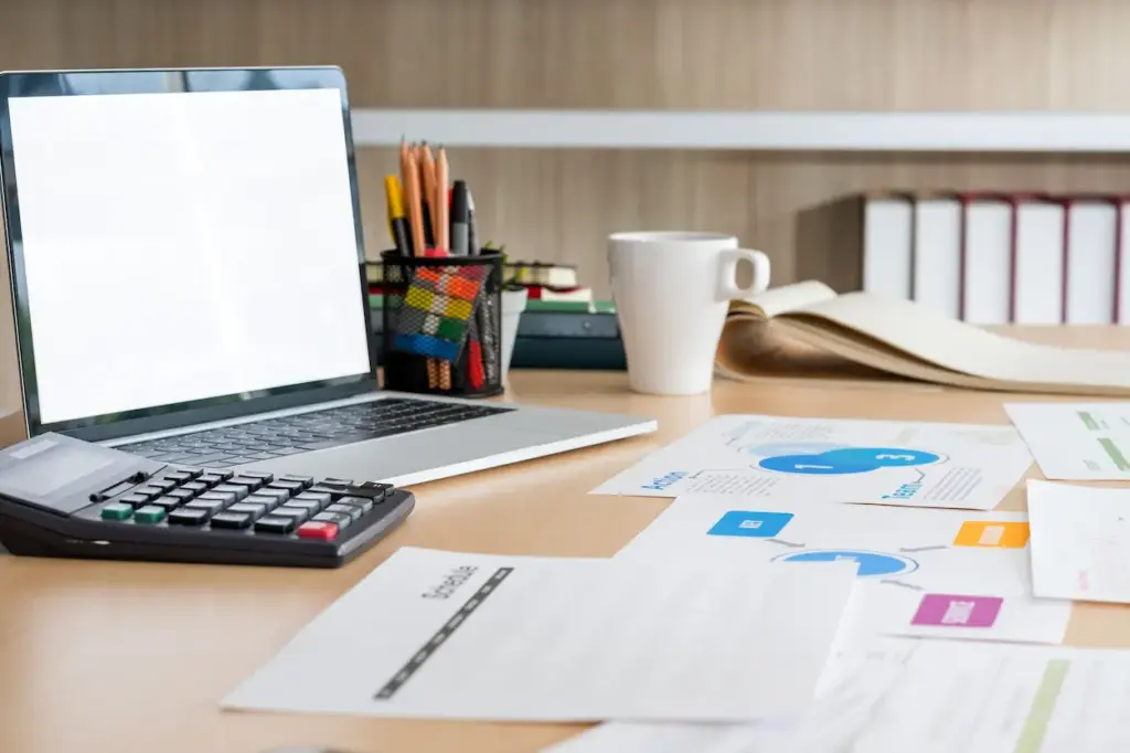 A desk with a laptop displaying a blank white screen, a calculator, a cup of pens, and business documents for a bookkeeping service in Tampa.
