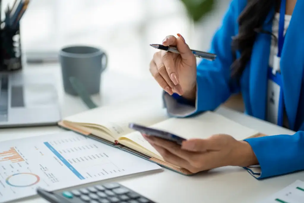 A person in a blue blazer holds a pen and smartphone while sitting at a desk with an open notebook and financial charts for food truck bookkeeping.
