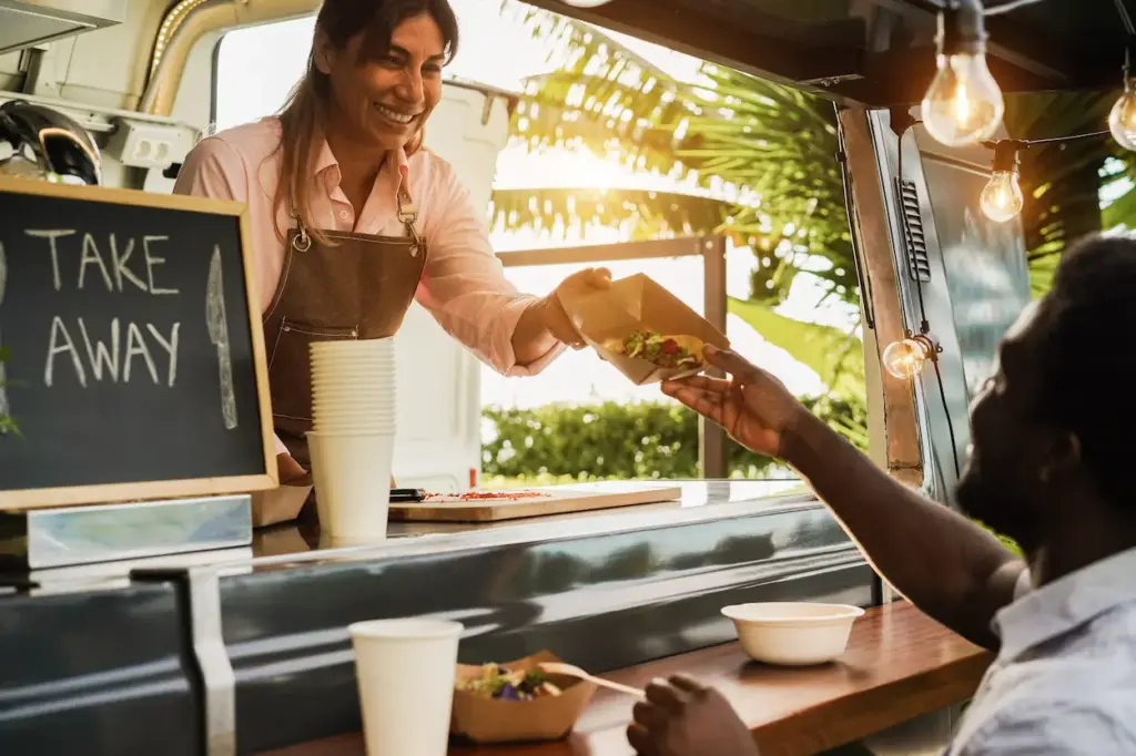 A food truck worker hands a taco in a cardboard tray to a customer during a shift involving food truck bookkeeping.