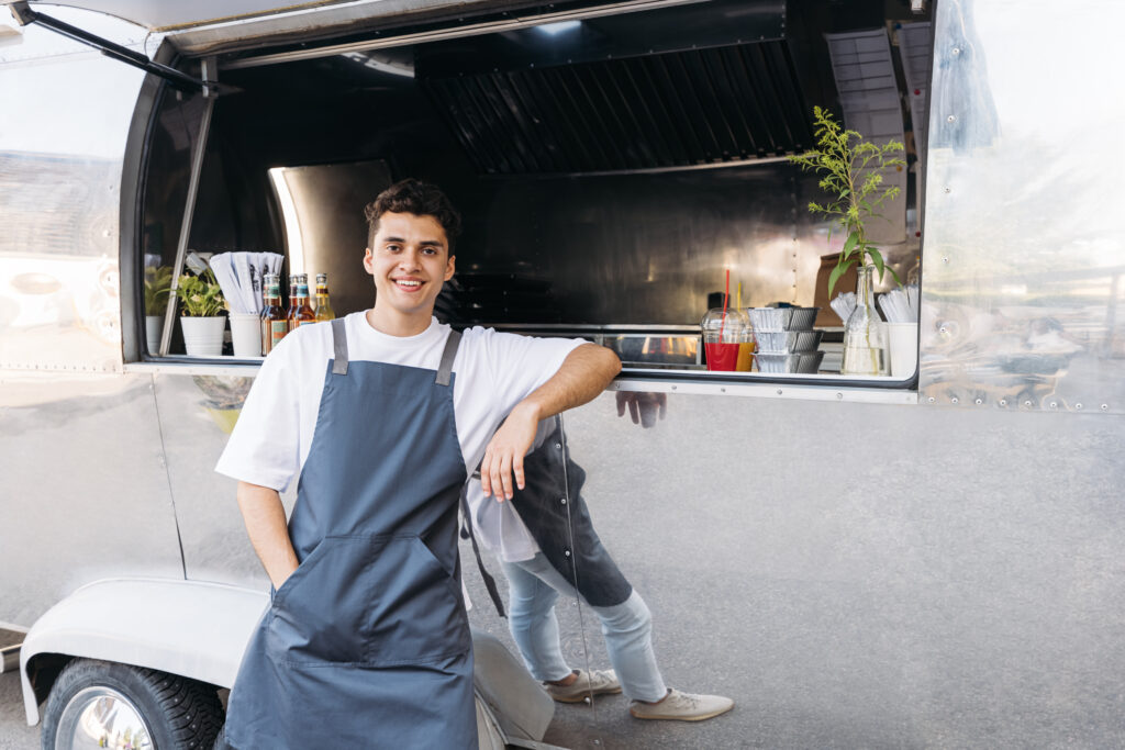 A smiling Kraken Bookkeeping employee in a white t-shirt and grey apron leans against the service window of a stainless steel food truck.