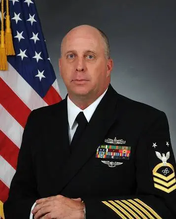 A U.S. Navy officer in a dress blue uniform with medals and insignia stands in front of an American flag, representing the precision required for construction bookkeeping.