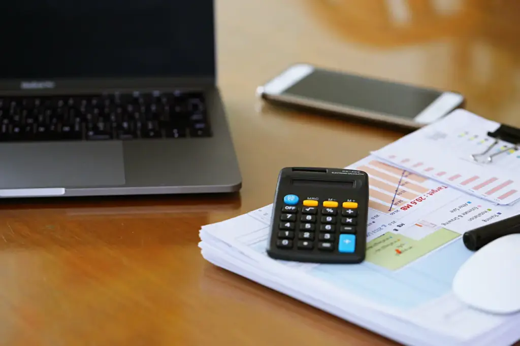 A laptop, smartphone, and black calculator resting on top of Kraken Bookkeeping financial documents on a wooden desk.